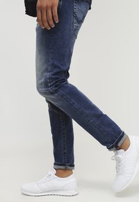 Jeans de mezclilla azul oscuro con efecto desvanecido, diseño ajustado y puños enrollados. Combinados con zapatillas deportivas blancas con tres franjas.