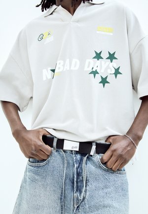 Overmaat lichtgrijze polo met een kraag en grafische print; groene sterren en de tekst "N BAD DAYS." Zwarte riem gecombineerd met lichtblauwe spijkerbroek.