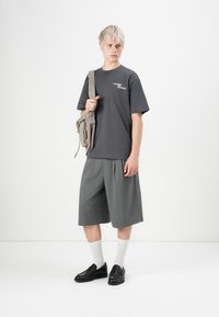 Junge Person mit kurzen blonden Haaren, die ein dunkelgraues T-Shirt, graue weite Shorts, weiße Socken, schwarze Schuhe trägt und eine beige Umhängetasche dabei hat.
