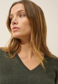 Sweter w serek w ciemnozielonym kolorze z wełny o miękkiej fakturze. Modelka ma brązowe włosy sięgające do ramion i neutralne tło.