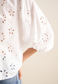 Blusa bianca in cotone con ricami floreali e motivi a taglio, maniche a sbuffo e orlo arrotondato, abbinata a pantaloni in denim.