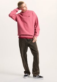 Felpa rosa con collo rotondo, maniche lunghe e piccolo logo. Abbinata a jeans baggy verdi scuri e sneaker nere con suola bianca.