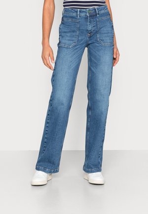 Personne portant un jean bleu droit avec de grandes poches plaquées à l'avant et des baskets blanches, debout devant un fond clair uni.