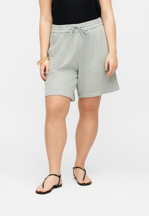 Nainen, jolla on vaaleanharmaat narusäätöiset shortsit, musta hihaton toppi, mustat sandaalit ja läpikuultava kultainen rannekoru vasemmassa ranteessa.