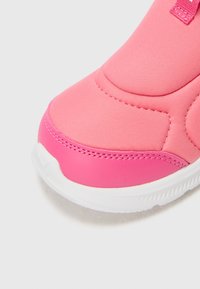 Zapatilla deportiva rosa con parte superior de textil suave, puntera redondeada, ribete elástico y suela de goma blanca. Los acentos incluyen costuras visibles.