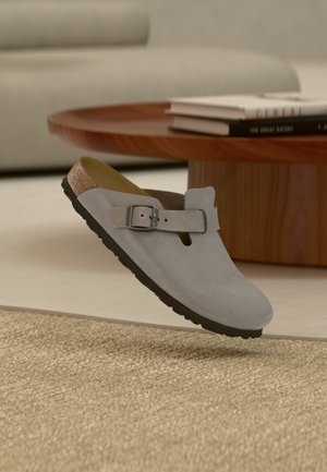 Sabot gris à enfiler unique avec boucle réglable flottant au-dessus d'un tapis beige texturé, table en bois et canapé en arrière-plan.