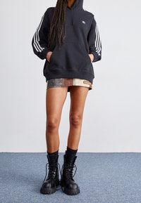 Sudadera negra con rayas blancas en las mangas y un logo, combinada con una falda corta de estampado metálico y botas de combate negras con cordones.