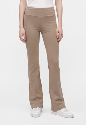 Άτομο που φοράει μπεζ καμπάνα παντελόνια και λευκά αθλητικά, στέκεται σε ένα απλό ανοιχτό φόντο.