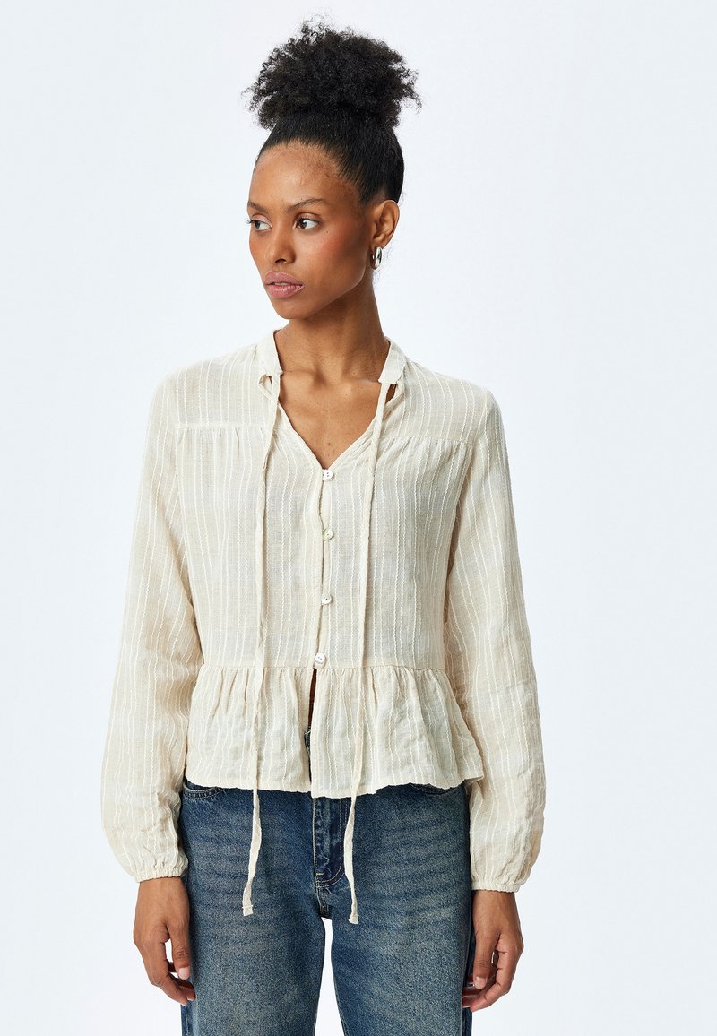 Blusa a bottoni beige chiaro con vita a peplum, righe verticali, maniche lunghe e dettagli con cordoncino al collo. Tessuto testurizzato.