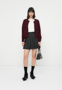 Bordeaux pletený cardigan s knoflíky, bílý top, skládaná černá sukně, černé lodičky na vysokém podpatku s mašlí a malá černá kabelka.