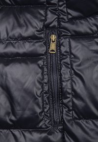 Primer plano de la tela de una chaqueta negra acolchada que presenta una cremallera vertical con un tirador de bronce etiquetado "NAPAPIJRI".