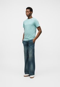 Άνδρας όρθιος με τα χέρια στις τσέπες, φοράει ανοιχτό μπλε μπλουζάκι, ξεβαμμένα τζιν με φαρδιά μπατζάκια και λευκά αθλητικά παπούτσια σε απλό λευκό φόντο.