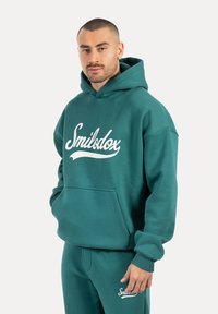 Sudadera de color teal con un bolsillo canguro, con el logo blanco de "Smilodox" en cursiva. Hecha de una tela suave con puños y dobladillo acanalados.