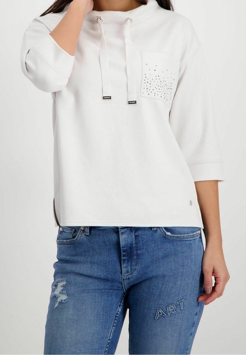 Pullover bianco con colletto alto, due cordini e tasca sul petto con borchie decorative. Abbinato a jeans blu strappati.