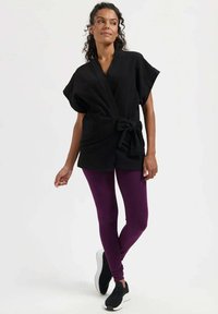 Schwarzes Wickeloberteil mit weiten Ärmeln und Binddetail, kombiniert mit violetten Leggings und schwarzen Sportschuhen vor neutralem Hintergrund.