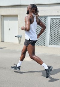 Czarne sportowe buty z teksturowanym wzorem, białe skarpetki oraz biała koszulka bez rękawów z delikatnymi wzorami, w zestawie z czarnymi szortami, na chodniku.