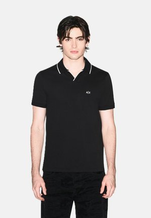 Giovane uomo con capelli scuri che indossa una polo nera a maniche corte con bordi bianchi e un piccolo logo A|X sul petto, in piedi davanti a uno sfondo neutro.