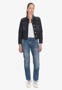 Veste en denim noire avec deux poches poitrine, fermetures par boutons argentés, débardeur blanc en dessous, associée à un jean bleu et des baskets claires.