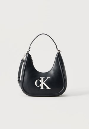 Czarna skórzana torba na ramię z srebrnym logo "cK", o zaokrąglonym kształcie, krótkim uchwytem i odpinanym dłuższym paskiem na jednolitym tle.
