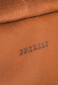 Bruin leer met een gladde textuur, voorzien van reliëf "BURKELY" branding. De kleur is warm en aards, met de nadruk op een natuurlijke afwerking.