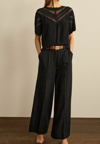 Blouse noire avec détails en dentelle sur les épaules, associée à un pantalon noir à jambes larges. Ceinture marron avec boucle géométrique. Sandales marron clair.