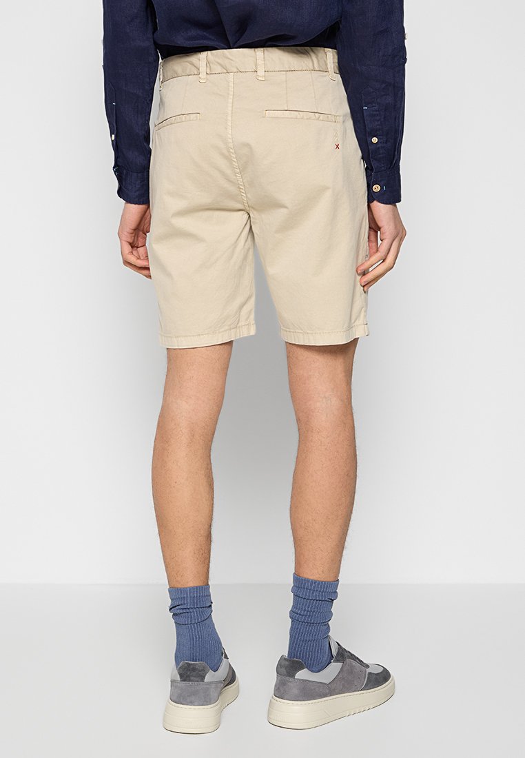 Beżowe bawełniane szorty o prostym kroju, z tylnymi kieszeniami i płaskim przodem. Noszone z niebieskimi skarpetkami w prążki i szarymi trampkami.