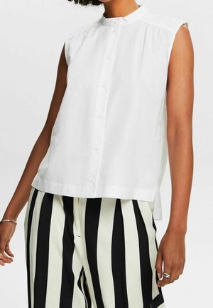 Chemise blanche sans manches à petits boutons et col montant, associée à un pantalon taille haute rayé vertical noir et blanc.