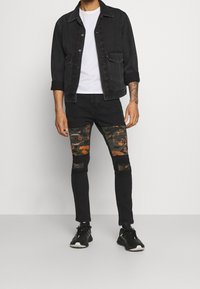 Chaqueta de mezclilla negra con cierre de botones, llevada sobre una camiseta blanca. Jeans negros ajustados con parches de rodilla en camuflaje naranja y verde y áreas desgastadas.