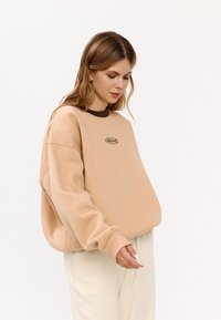 Felpa beige oversize con scollatura tonda e dettaglio marrone; presenta un logo ricamato al centro. Tessuto morbido e vestibilità rilassata.