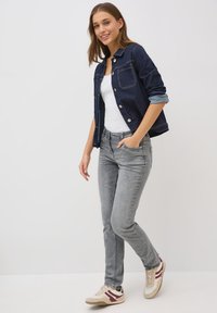 Donkerblauwe spijkerjacket, witte T-shirt, grijze jeans en beige sneakers met maroon accenten. Model staat met handen in de zakken en opgerolde mouwen.