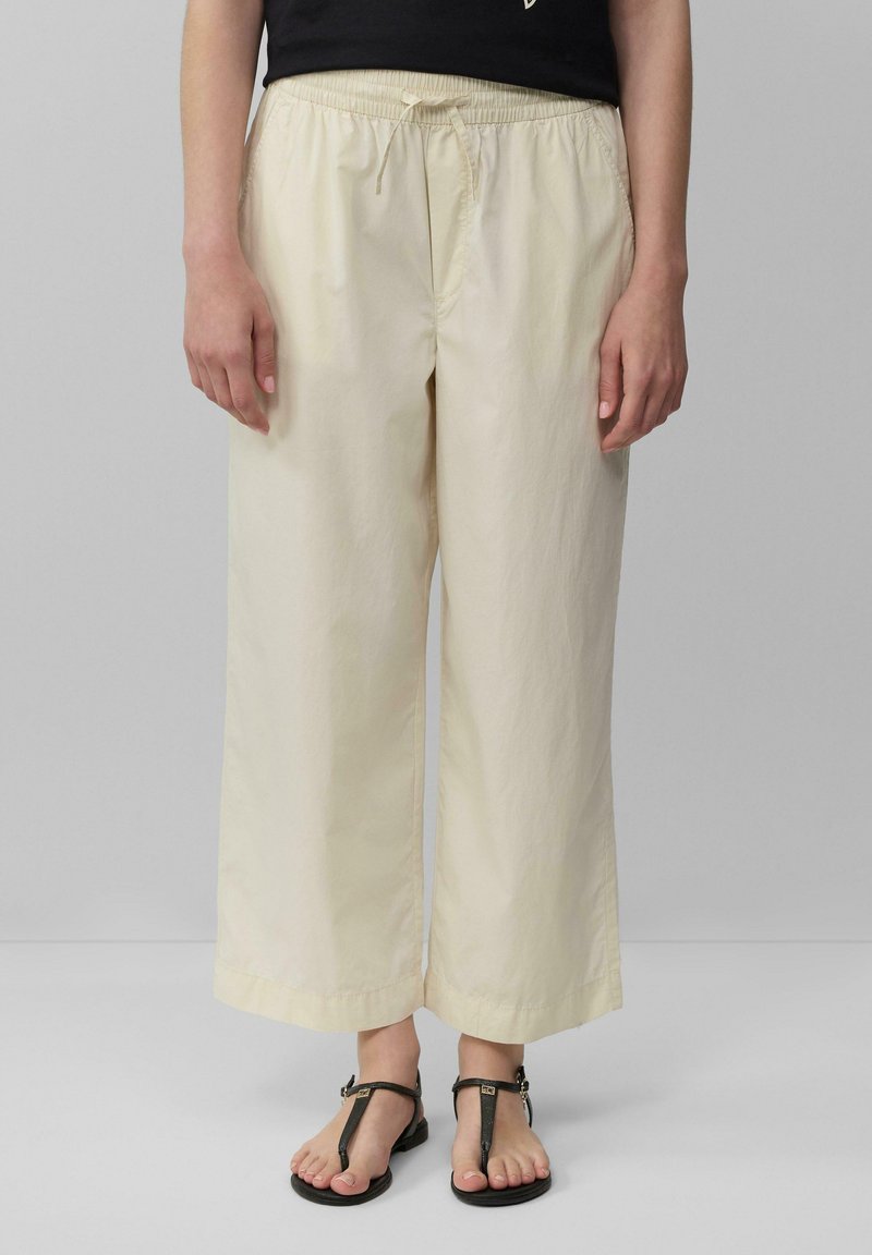 Helle beige weit geschnittene Hose mit elastischem Bund und Kordelzug, aus einem glatten, leichten Stoff. Kombiniert mit schwarzen Sandalen.