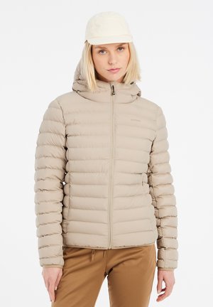 Jong persoon die een beige gewatteerd jack met capuchon, een beige pet en een bruine broek draagt, staand tegen een effen witte achtergrond.