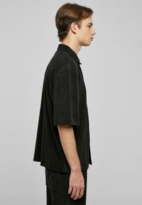 Camisa negra de manga corta con un ajuste holgado, cuello con solapa y textura suave, exhibida en un modelo de perfil.