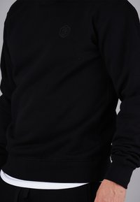 Black sweatshirt featuring a crew neck, ribbed cuffs and hem, and a small black logo on the chest. Made from a soft, textured fabric.