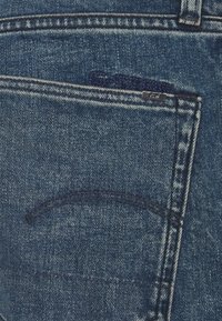 Modré džíny z denimu se standardní zadní kapsou, s malou značkovou nálepkou a viditelným švem v kontrastní tmavě modré barvě.