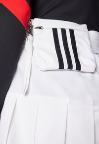 Μαύρη και κόκκινη μπλούζα με μια λευκή πλισέ φούστα. Λευκή τσάντα με φερμουάρ στη μέση, που διαθέτει τρεις μαύρες ρίγες.