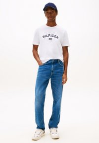 Witte T-shirt met "HILFIGER" logo, blauwe denim jeans, marineblauwe honkbalpet en witte sneakers. Casual pasvorm met klassieke ontwerp elementen.