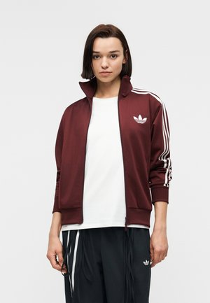 Jauna moteris dėvi bordo spalvos Adidas sportinį švarkelį su baltomis juostelėmis ir logotipu, baltą marškinėlį ir juodas Adidas kelnes su baltomis juostelėmis.