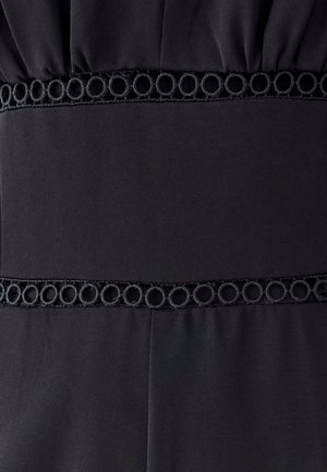 Schwarzes Stoffpaneel mit strukturiertem kreisförmigem Stickbesatz an den oberen und unteren Kanten, auf plissiertem Stoffhintergrund genäht.