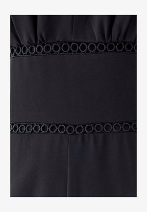 Schwarzes Stoffpaneel mit strukturiertem kreisförmigem Stickbesatz an den oberen und unteren Kanten, auf plissiertem Stoffhintergrund genäht.