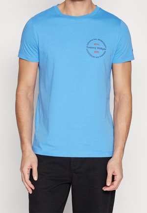 Mann trägt hellblaues Tommy Hilfiger T-Shirt mit schwarzen Hosen, kleines rundes Logo auf der linken Brust mit Text und Zahlen.