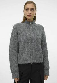 Grå zip-up genser med stående krage, ribbete tekstur og vide ermer, kombinert med svarte bukser. Mykt, varmt materiale.