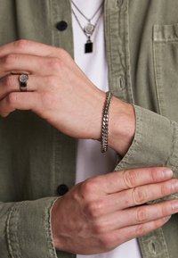 Bracelet en chaîne métallique au poignet ; couleur argentée avec une texture lisse. Une bague assortie au doigt présente un centre rond texturé.