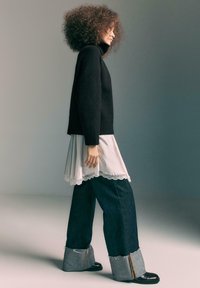 Maglione nero lavorato a maglia, sovrapposto a un vestito leggero con bordi in pizzo, abbinato a jeans di jeans scuri a gamba larga con orlo raw e polsini a contrasto.