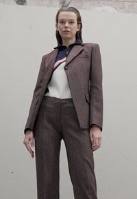 Γυναίκα που στέκεται μπροστά σε μια απλή τοιχογραφία, φορώντας ένα καφέ υφασμάτινο κοστούμι με στενή σακάκι και φαρδιά παντελόνια, και ένα τοπ με μαύρο και λευκό μοτίβο.