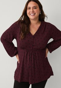 Smilende kvinde med langt brunt hår iført en bordeaux blouse med sorte prikker og sorte bukser, hænderne hvilende på hoften.