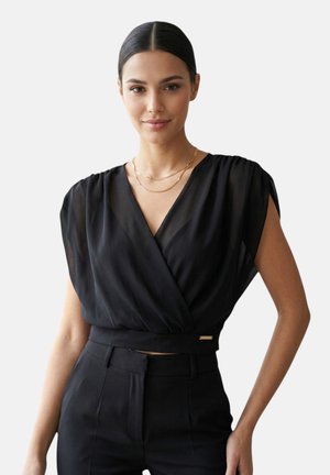 Donna con capelli scuri raccolti indossa un top nero senza maniche a portafoglio e pantaloni neri a vita alta con due collane d'oro su uno sfondo semplice.