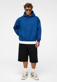 Sweatshirt à capuche bleue avec une poche devant et des cordons de serrage, associé à un short noir et des baskets de sport dans des tons neutres.