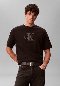 Νέος άνδρας που φορά μαύρο t-shirt Calvin Klein με λογότυπο, μαύρο τζιν, μαύρη ζώνη και ασημένιο βραχιόλι, στέκεται μπροστά σε απλό γκρι φόντο.