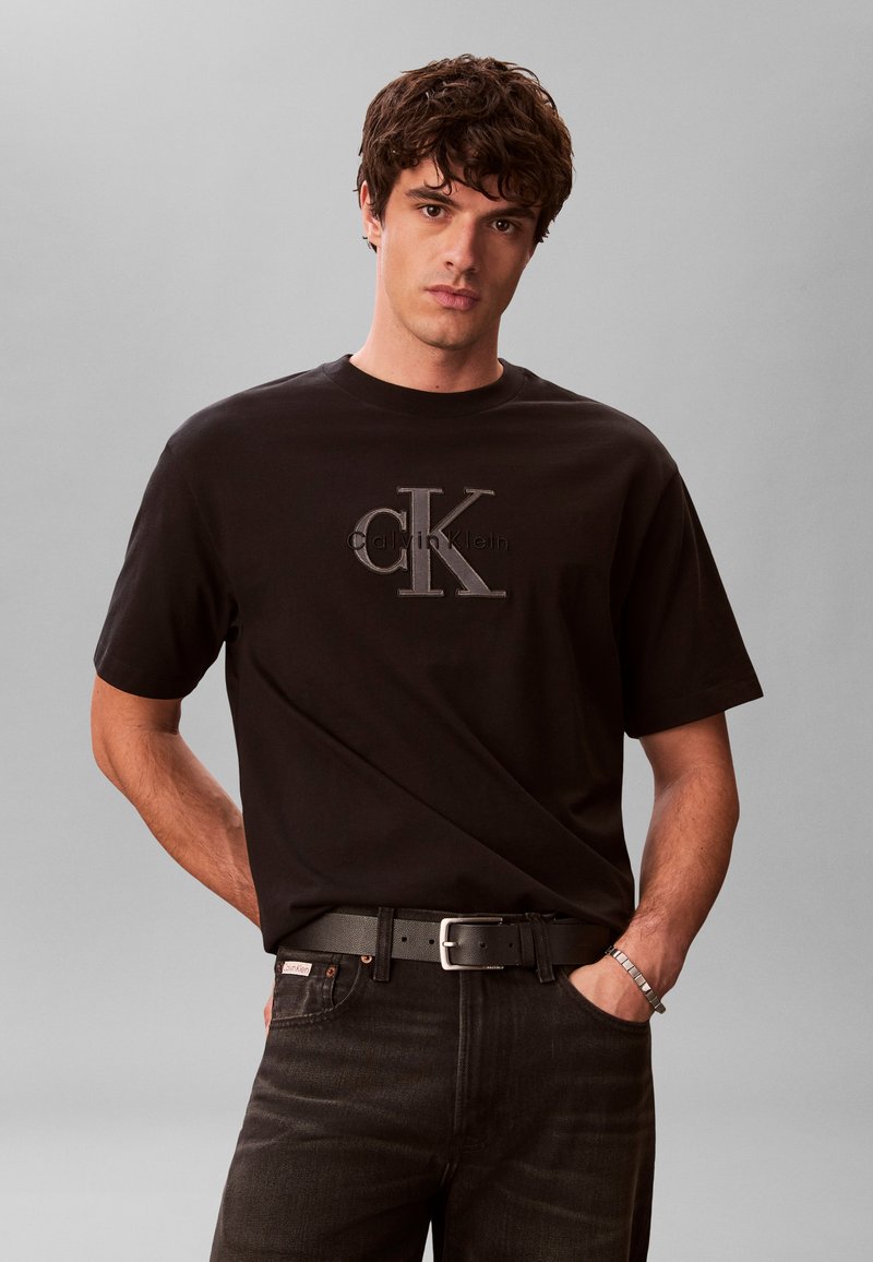 Νέος άνδρας που φορά μαύρο t-shirt Calvin Klein με λογότυπο, μαύρο τζιν, μαύρη ζώνη και ασημένιο βραχιόλι, στέκεται μπροστά σε απλό γκρι φόντο.